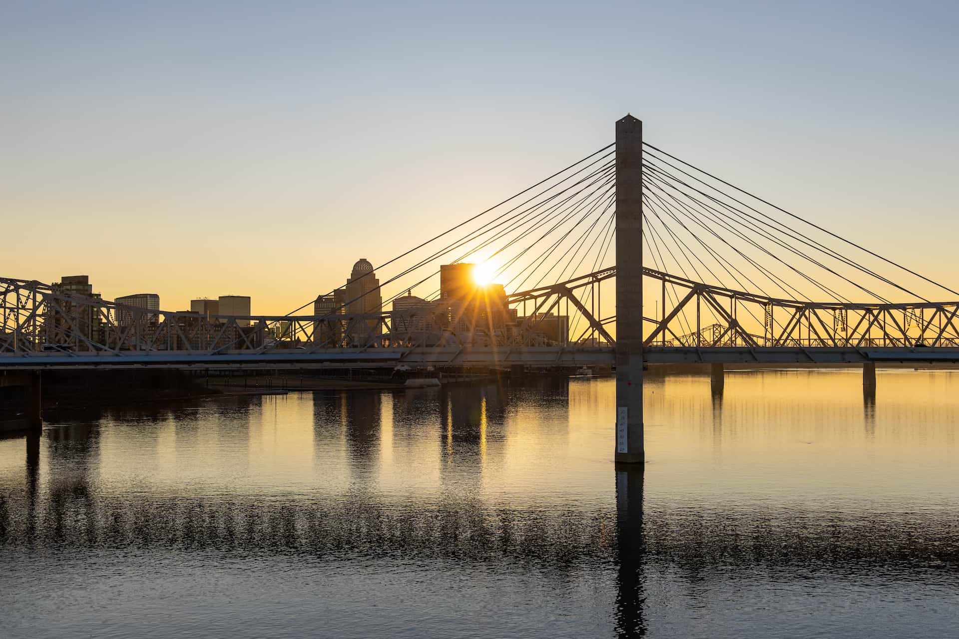 A large bridge running over a wide river in a city in Kentucky, with several tall buildings seen far off in the background.